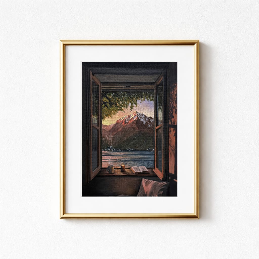A framed print showing a scenic view through an open window. The artwork features a mountain range, a lake, and a candle, book, and mug on the windowsill. The frame is gold.